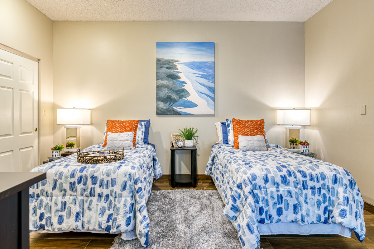 A cozy bedroom with two twin beds, decorated in blue and orange tones, featuring a beach painting and bedside lamps.