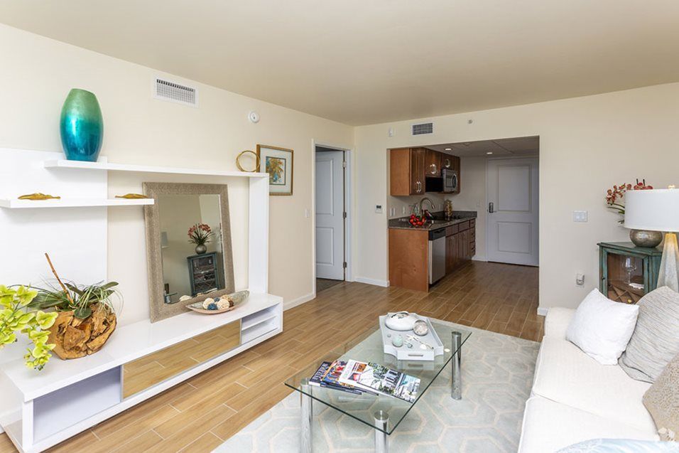 A modern apartment living room with wood flooring, a glass coffee table, and a view into the kitchen.