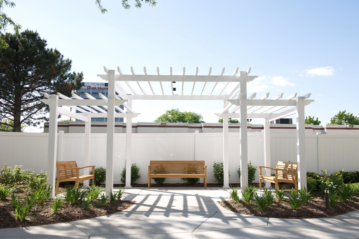 A serene garden setting with a white pergola, wooden benches, and a paved pathway under a clear sky.