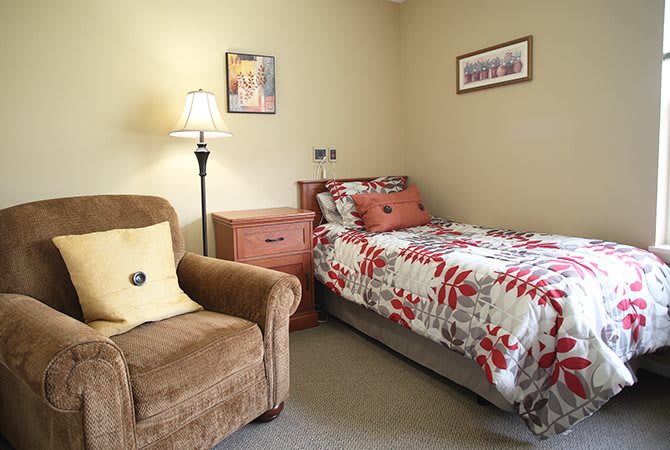 A cozy bedroom with a single bed and an armchair, complemented by a lamp and decorative pillows.