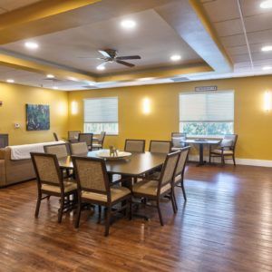 A spacious dining room with wooden floors, tables, and chairs, featuring large windows and warm yellow walls.