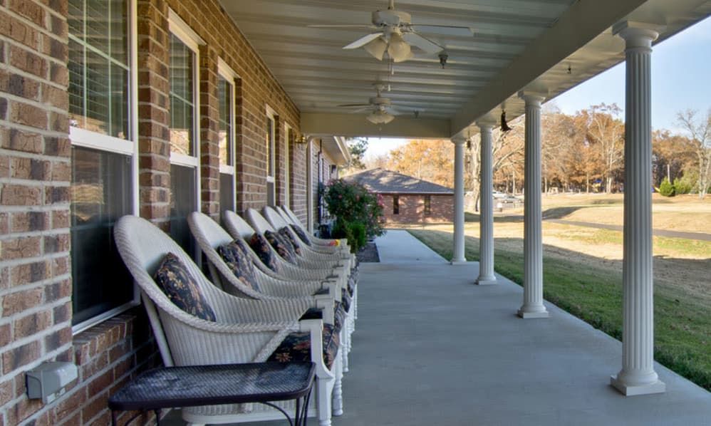 A row of white wicker chairs lined up on a covered porch with ceiling fans overhead.