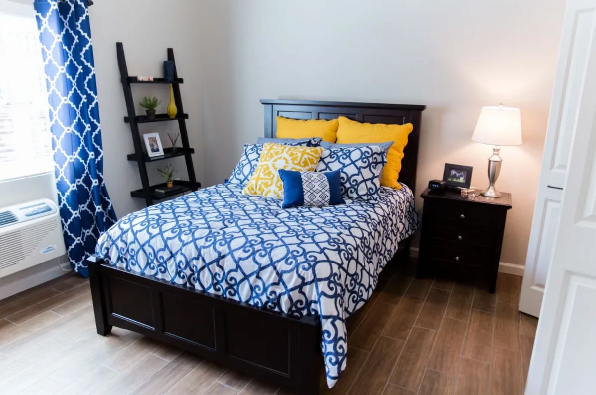 A well-decorated bedroom with a neatly made bed featuring blue and yellow pillows, alongside a lamp and decorative shelves.