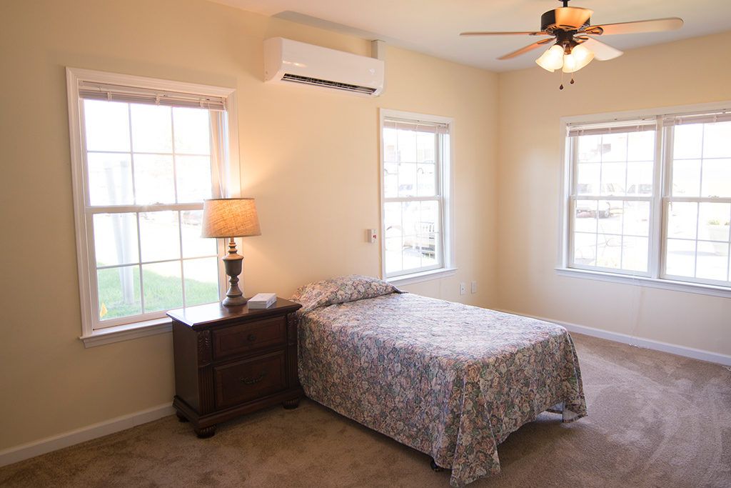 A cozy bedroom with a single bed, nightstand, and lamp, featuring an air conditioner and ceiling fan.