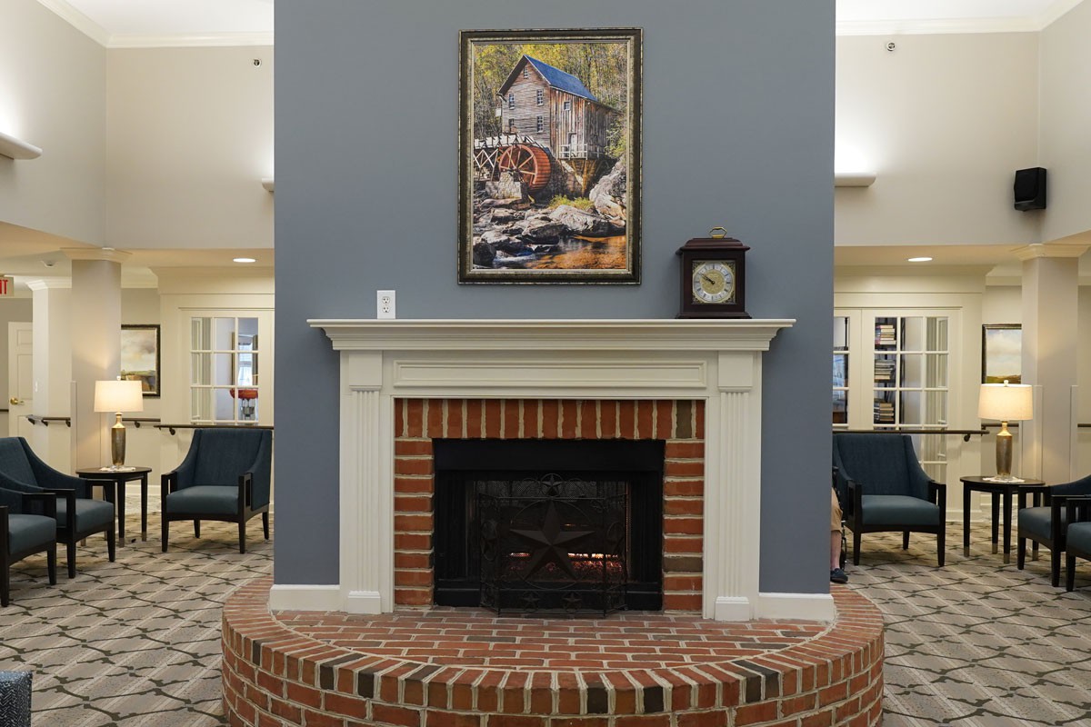 A cozy room featuring a brick fireplace with a painting and clock on the mantel, surrounded by comfortable chairs on a patterned carpet.