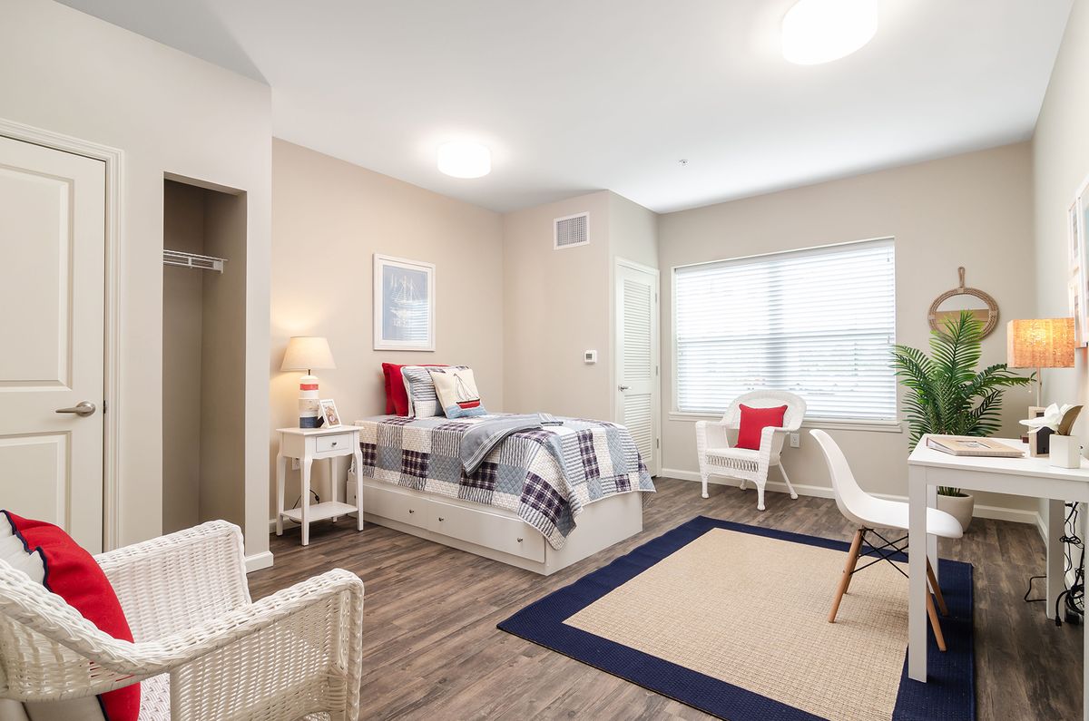 A modern bedroom featuring a bed with patterned bedding, two chairs, a small table lamp, and a large window for natural light.