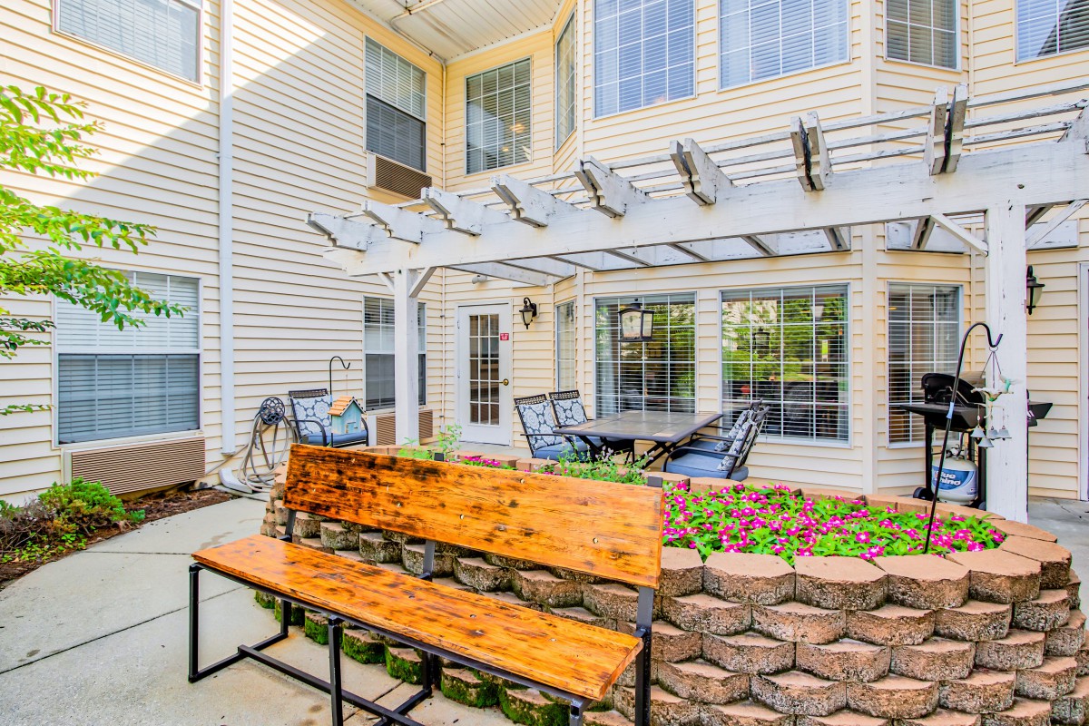 A cozy outdoor patio with a wooden bench, pergola, and seating area surrounded by plants.
