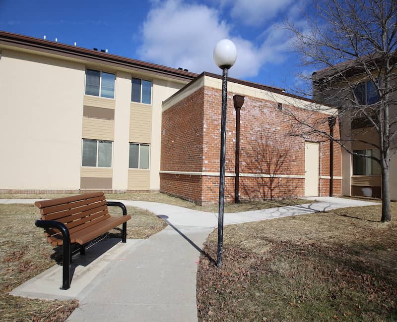 A courtyard with a bench and lamppost alongside a brick and stucco building under a partly cloudy sky.