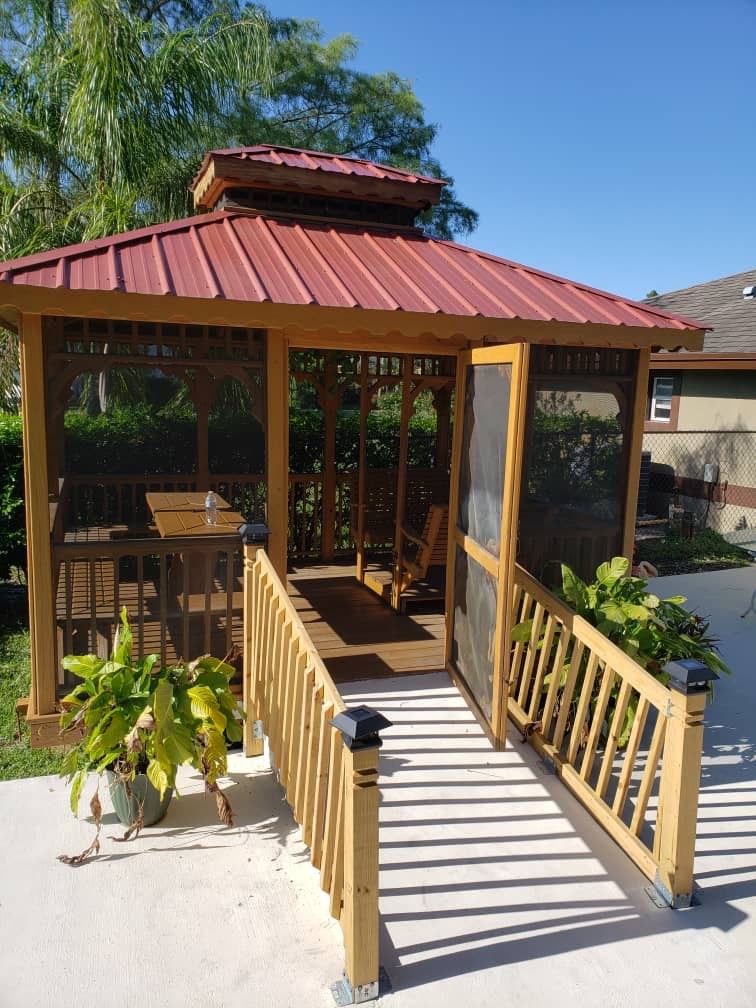 A cozy wooden gazebo with a red roof in a garden setting, surrounded by plants.
