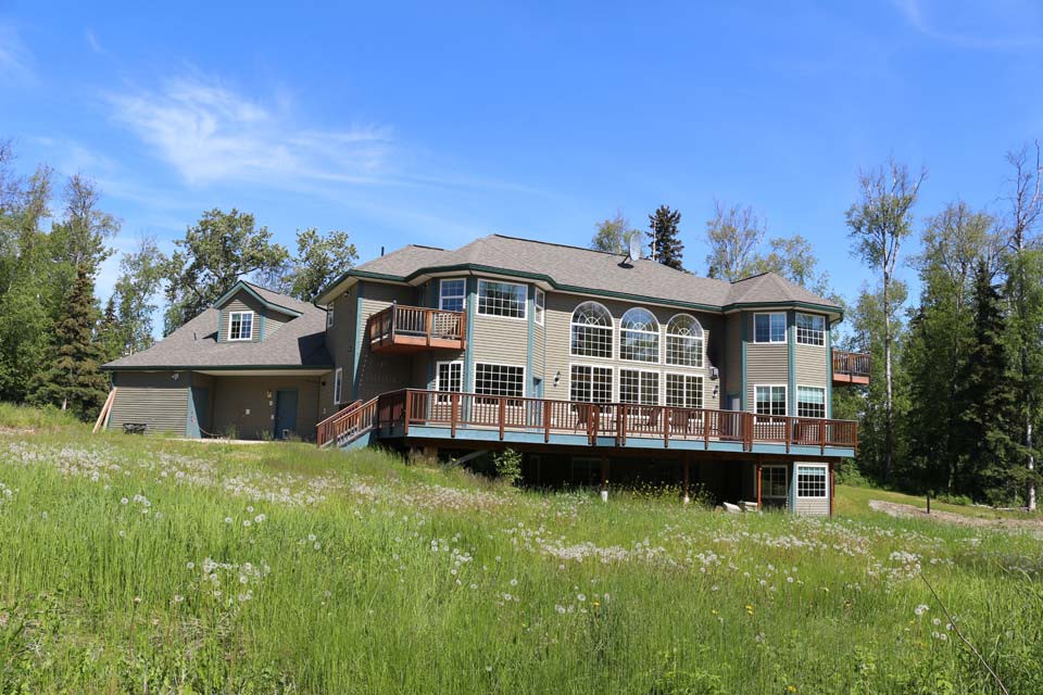 A large modern house with a spacious deck, surrounded by greenery and under a clear blue sky.