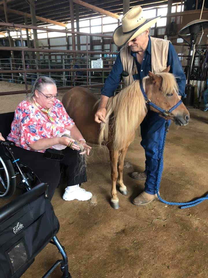 A woman in a wheelchair interacts with a pony, assisted by a man wearing a cowboy hat.