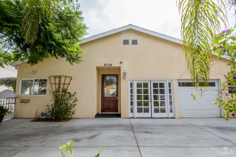The image shows the exterior of a beige-colored house with a driveway, wooden front door, and white garage doors.