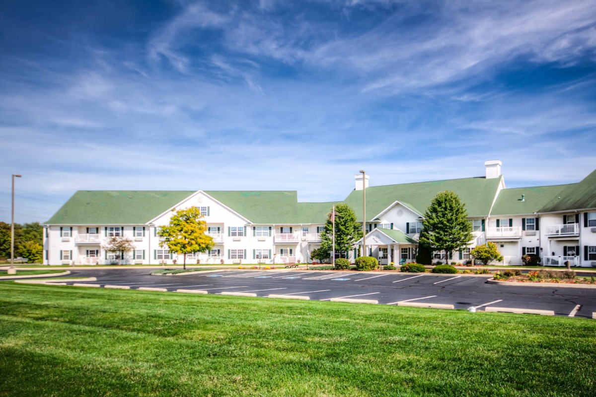 A large building with a green roof surrounded by trees and a parking lot under a blue sky.