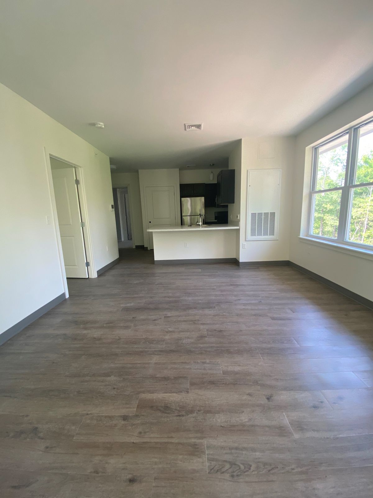 Spacious empty living room with wooden floors and a view into the kitchen, illuminated by natural light from a large window.