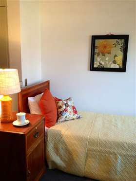 A cozy bedroom with a nightstand and lamp beside a neatly made bed, featuring decorative pillows and a framed painting on the wall.