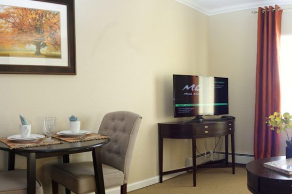 A cozy living room with a TV on a stand, a small round table with two chairs, and a framed picture on the wall.