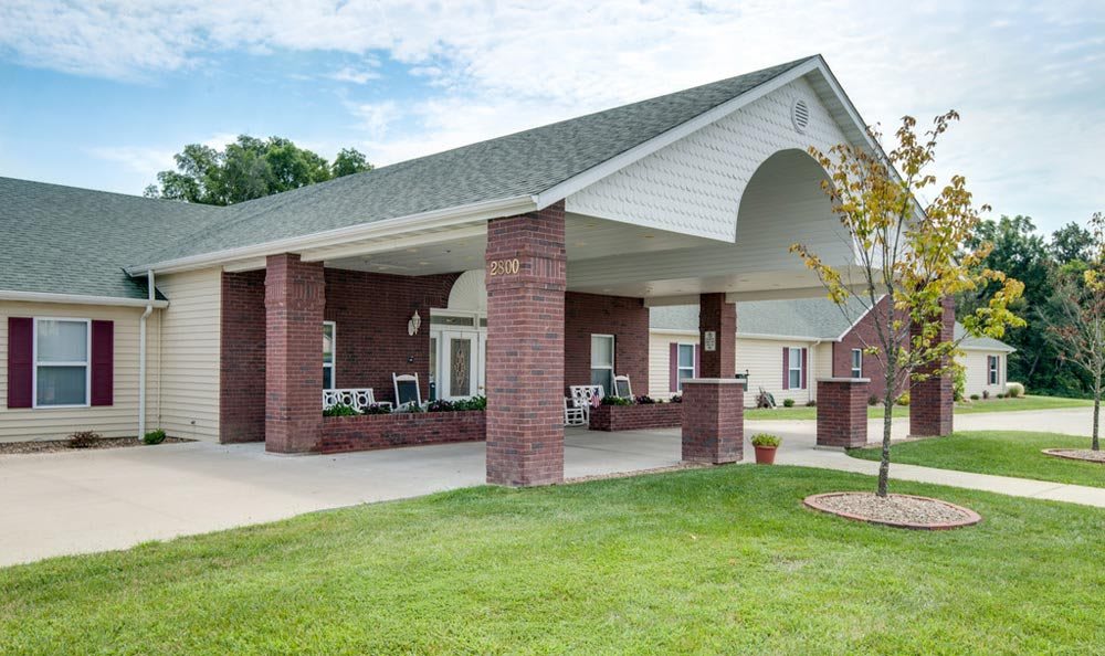 A building entrance with a covered area supported by brick columns, set in a landscaped outdoor environment.