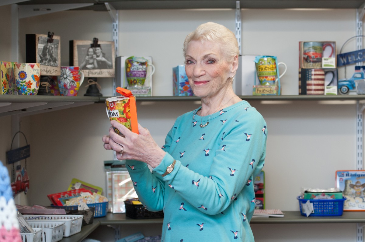 A woman smiling and holding an item in front of a shelf filled with colorful mugs.