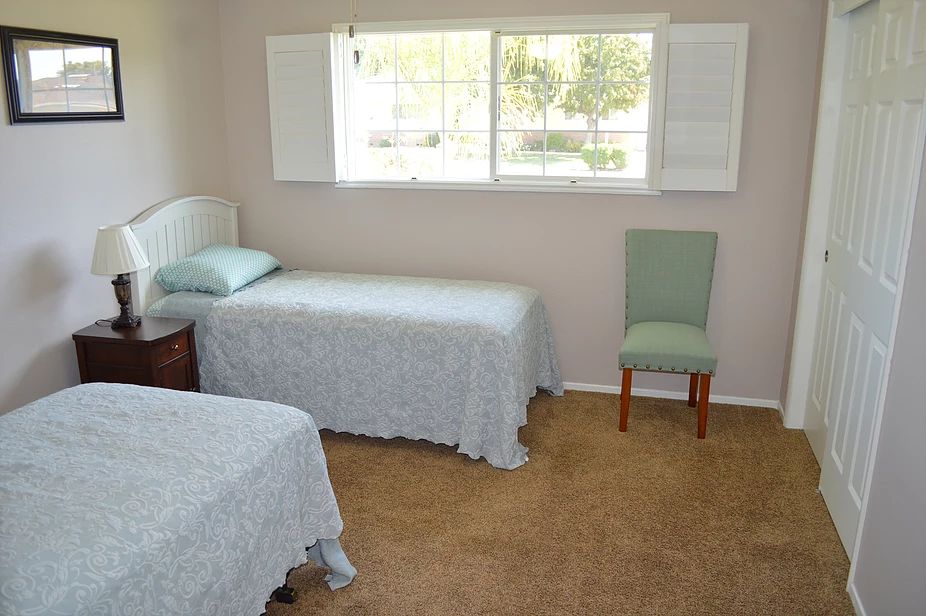 A simple bedroom with two twin beds, a chair, and a window, featuring light-colored bedding.