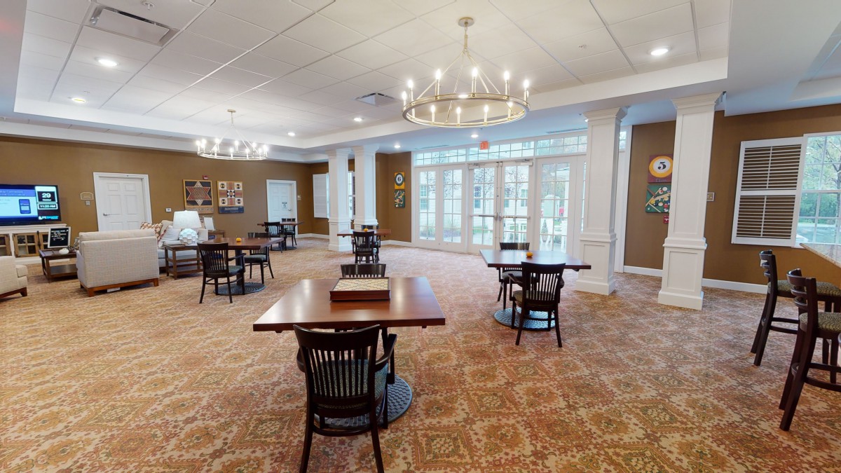 A spacious dining area with elegant chandeliers, wooden tables, and chairs arranged on a patterned carpet, complemented by large windows for natural light.
