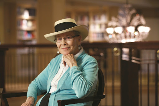An elderly woman wearing a stylish hat and a turquoise cardigan, smiling while seated indoors.