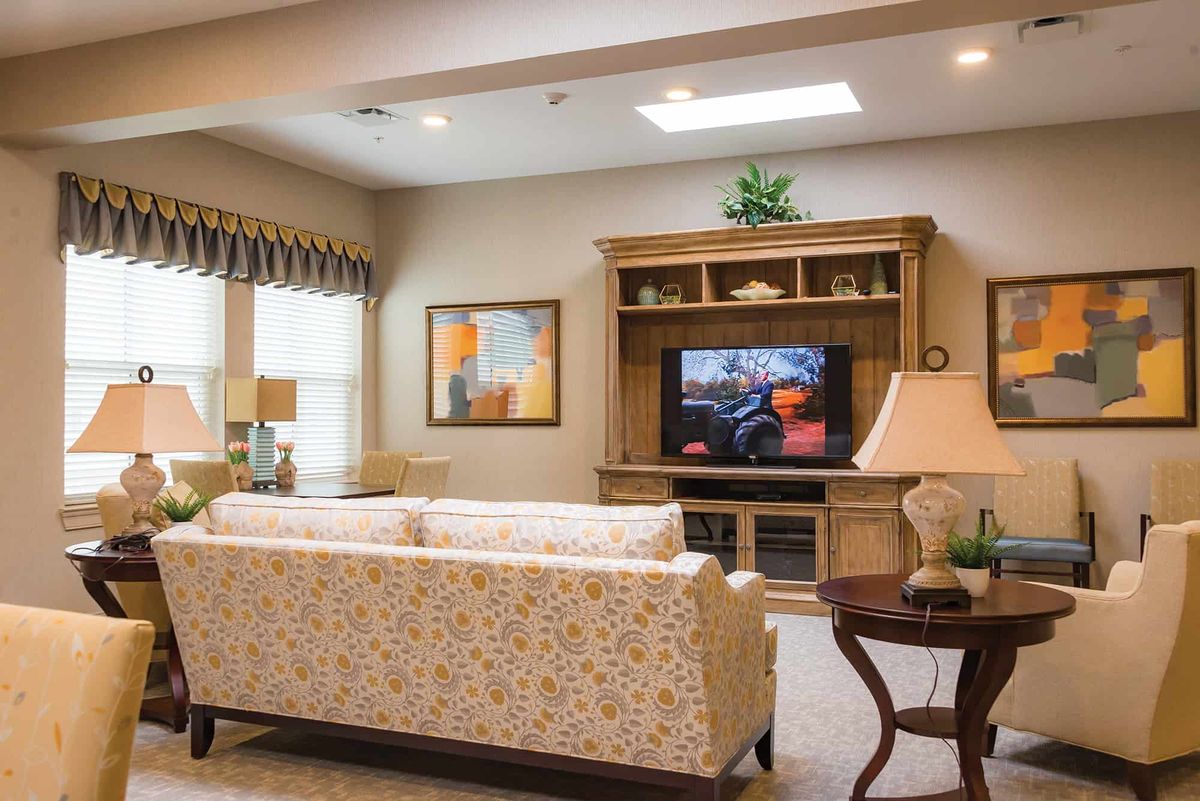 A cozy living room with a sofa facing a television, decorated with artwork and illuminated by natural light from the windows.