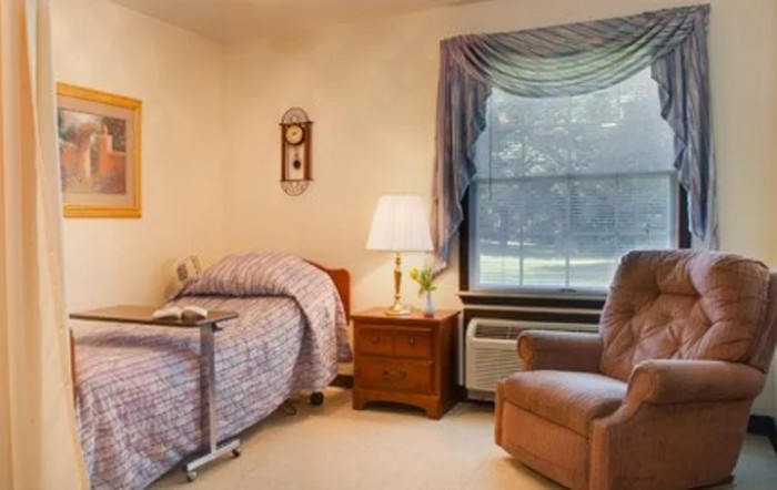 A cozy bedroom with a single bed, bedside table, lamp, and an armchair near a large window.