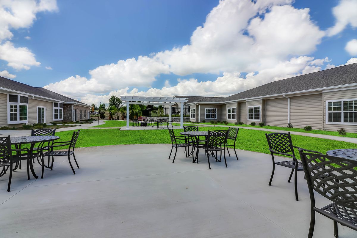 A spacious outdoor patio area with tables and chairs, surrounded by grass and buildings under a partly cloudy sky.
