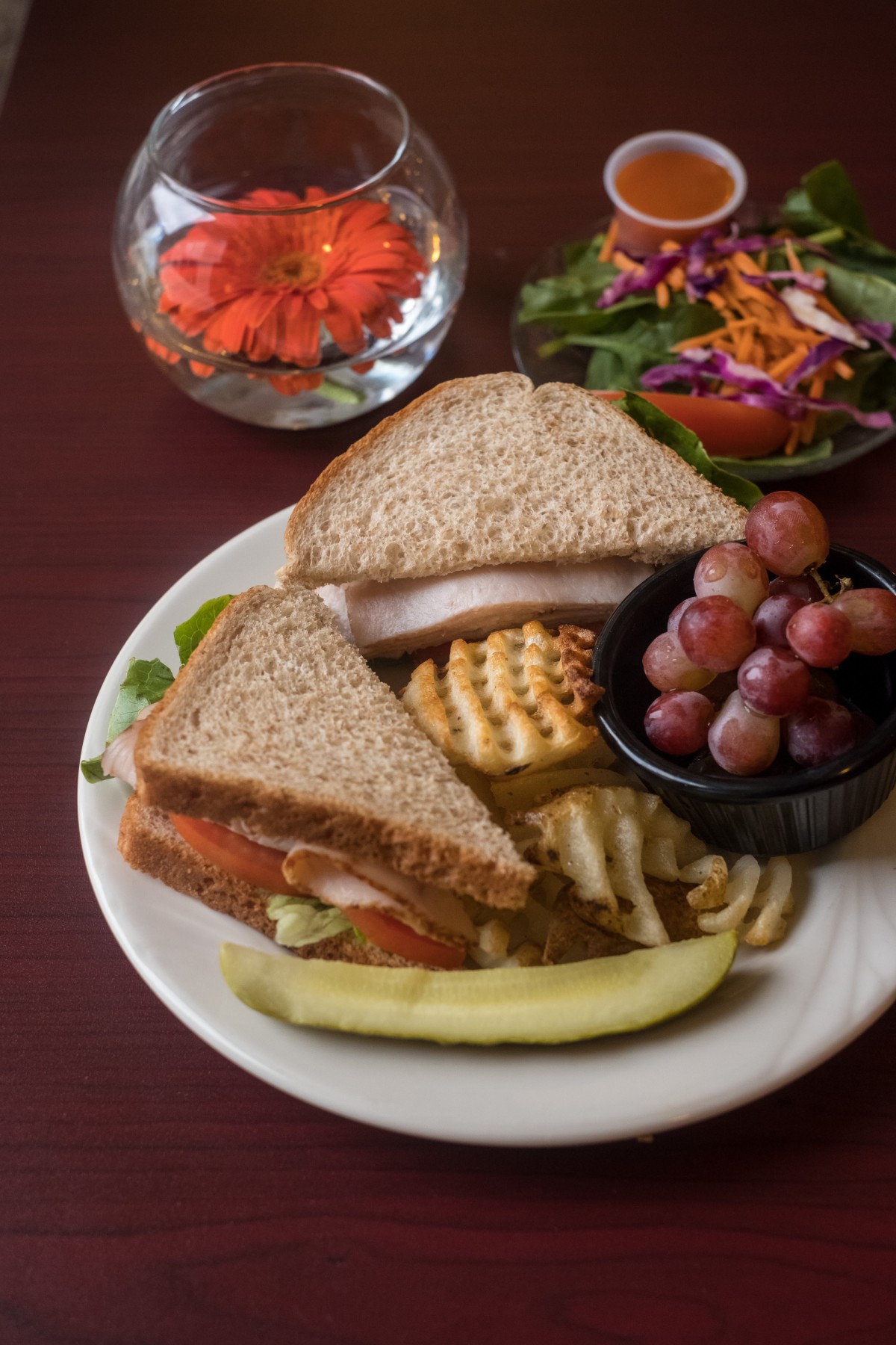 A plate featuring a sandwich with whole wheat bread, accompanied by grapes, crackers, and a pickle slice, with a side salad in the background.