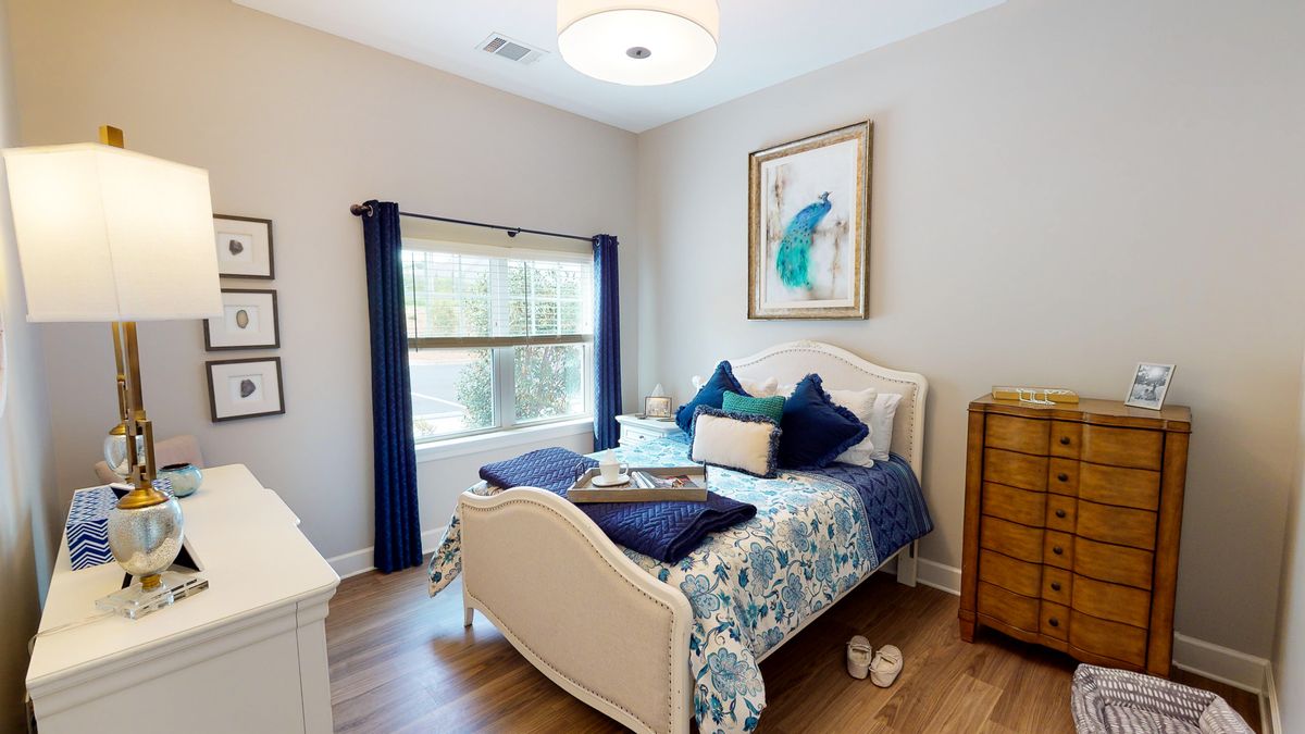 A cozy bedroom featuring a neatly made bed with blue-themed cushions, a framed artwork on the wall, and natural light streaming through a window with dark curtains.