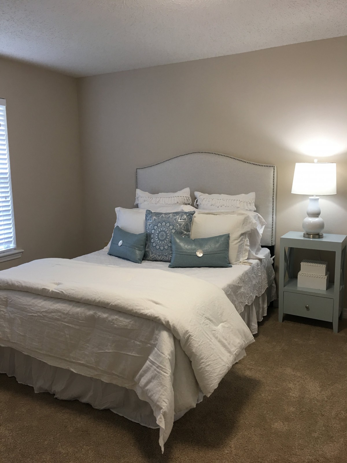 A cozy bedroom with a neatly made bed, featuring multiple pillows and a lit lamp on the nightstand.