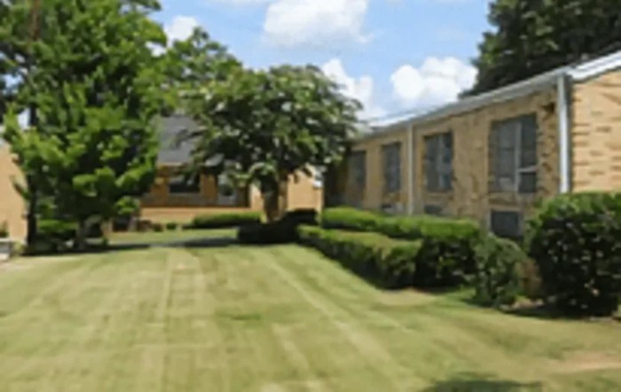 A well-maintained lawn with trees and bushes in front of a brick building under a partly cloudy sky.
