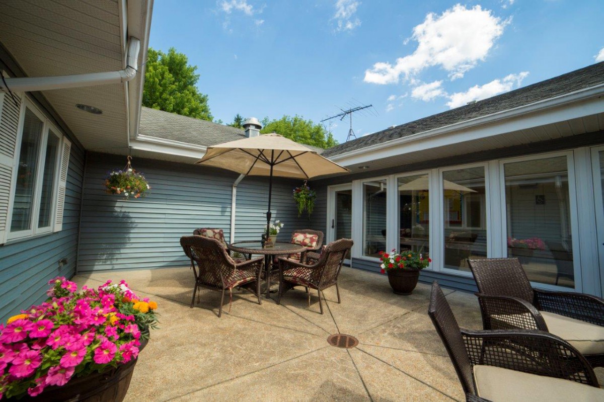 A sunny patio with outdoor furniture and a large umbrella surrounded by flowers.