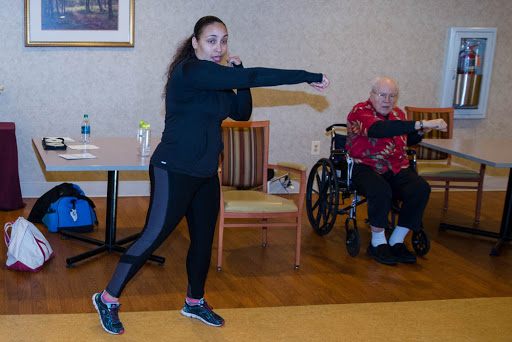 A fitness instructor demonstrates a workout move in a senior center, while an elderly participant engages from a wheelchair.