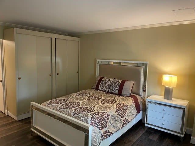 A cozy bedroom featuring a bed with patterned bedding, a white nightstand with a lamp, and a wardrobe.