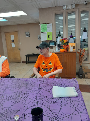 A festive Halloween gathering at a senior center, featuring residents dressed in bright orange shirts with pumpkin designs.