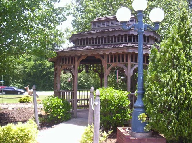 A wooden gazebo surrounded by bushes and a pathway, with a lamp post nearby.