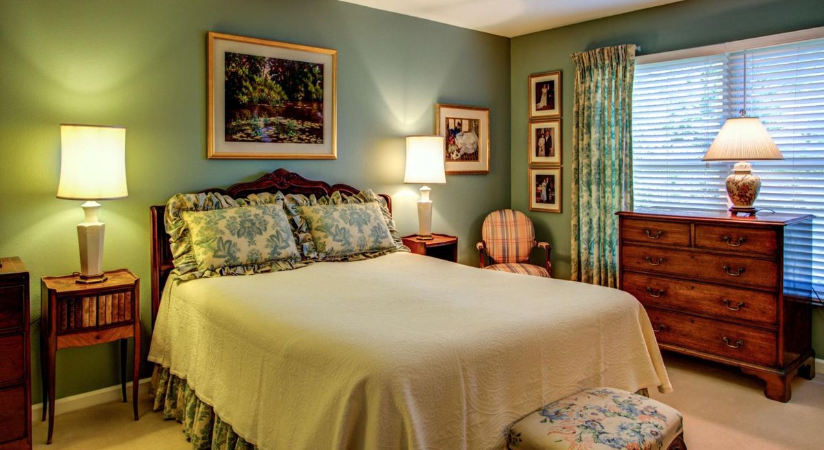 A traditionally styled bedroom with a neatly made bed, lamp, and framed artwork.