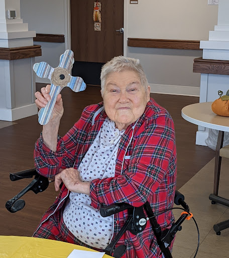 An elderly woman in a plaid jacket smiles while holding a cross, sitting in her wheelchair indoors.