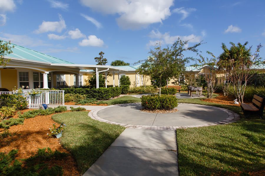 A beautifully landscaped garden with a circular pathway and surrounding buildings under a blue sky with clouds.