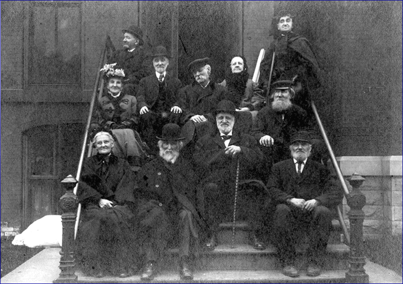 A historical black and white photo of a group of people in formal attire sitting on a staircase.