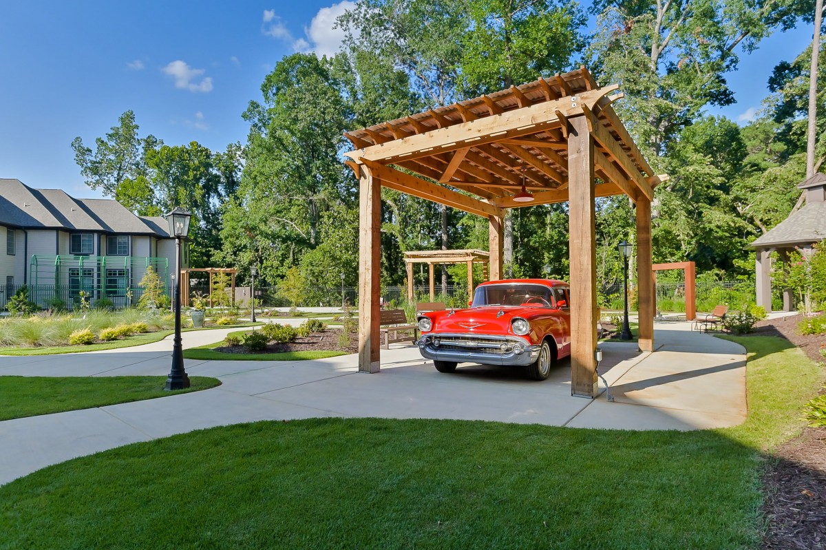 A vintage red car parked under a wooden carport surrounded by greenery and trees.