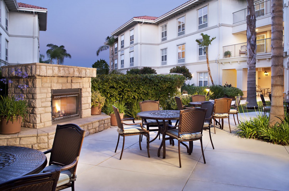 An inviting outdoor patio area with seating and a fireplace set against a backdrop of a well-lit building during the evening.