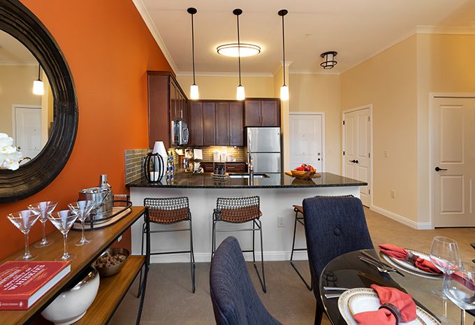 A modern kitchen with stainless steel appliances and an adjacent dining area, featuring an orange accent wall and stylish hanging lights.