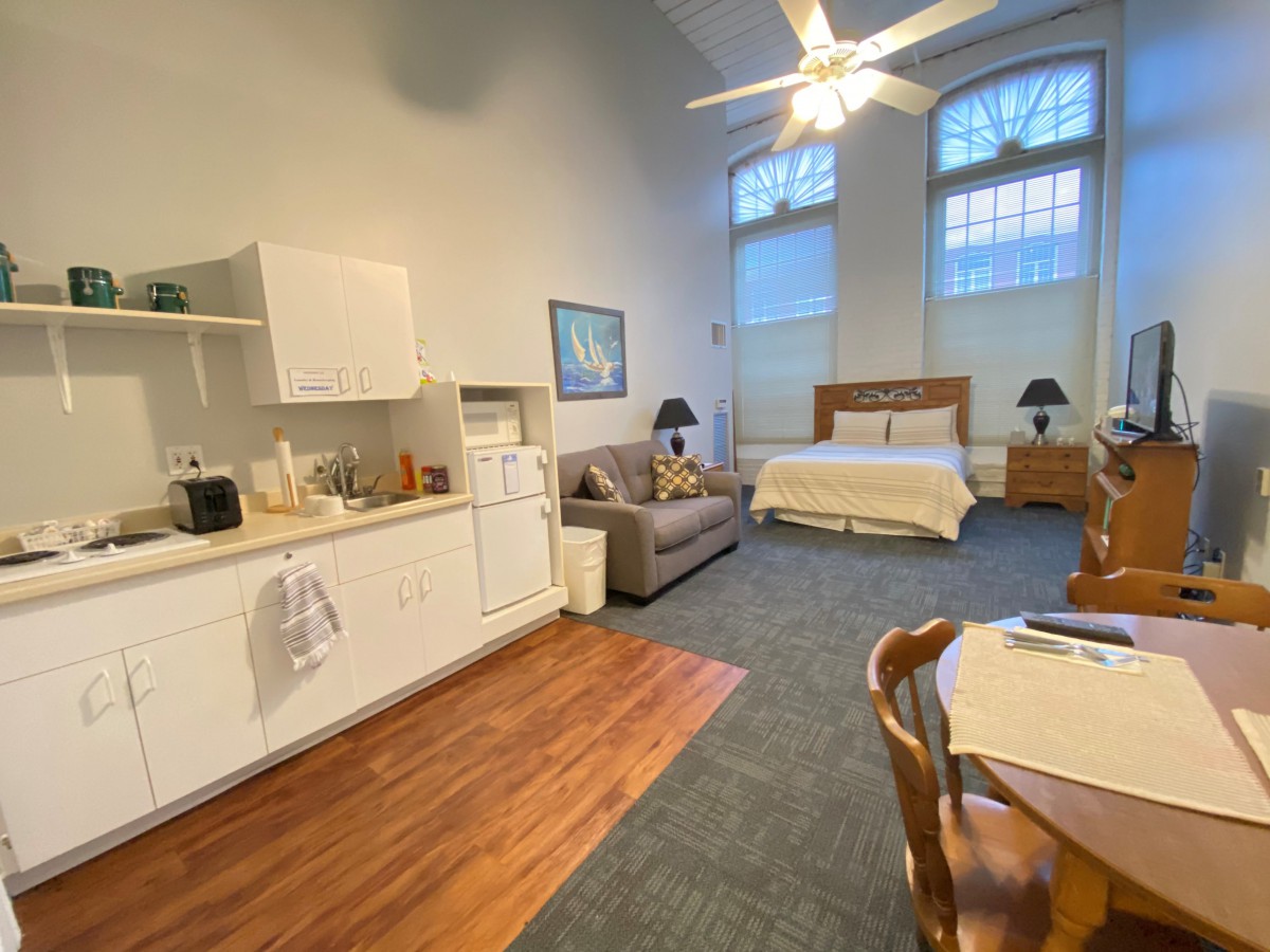 A cozy studio apartment featuring a kitchenette, living area, and bed with a ceiling fan above.