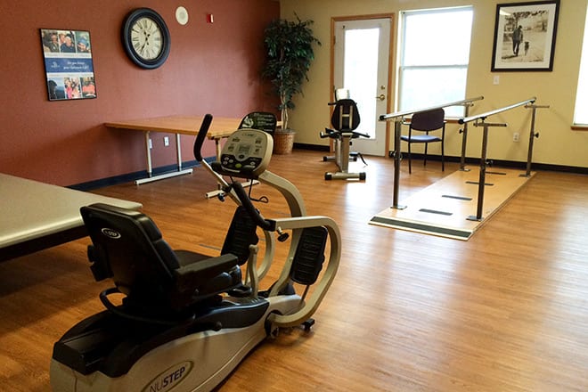 A rehabilitation exercise room featuring fitness equipment including a stationary bike, with wood flooring and natural light.