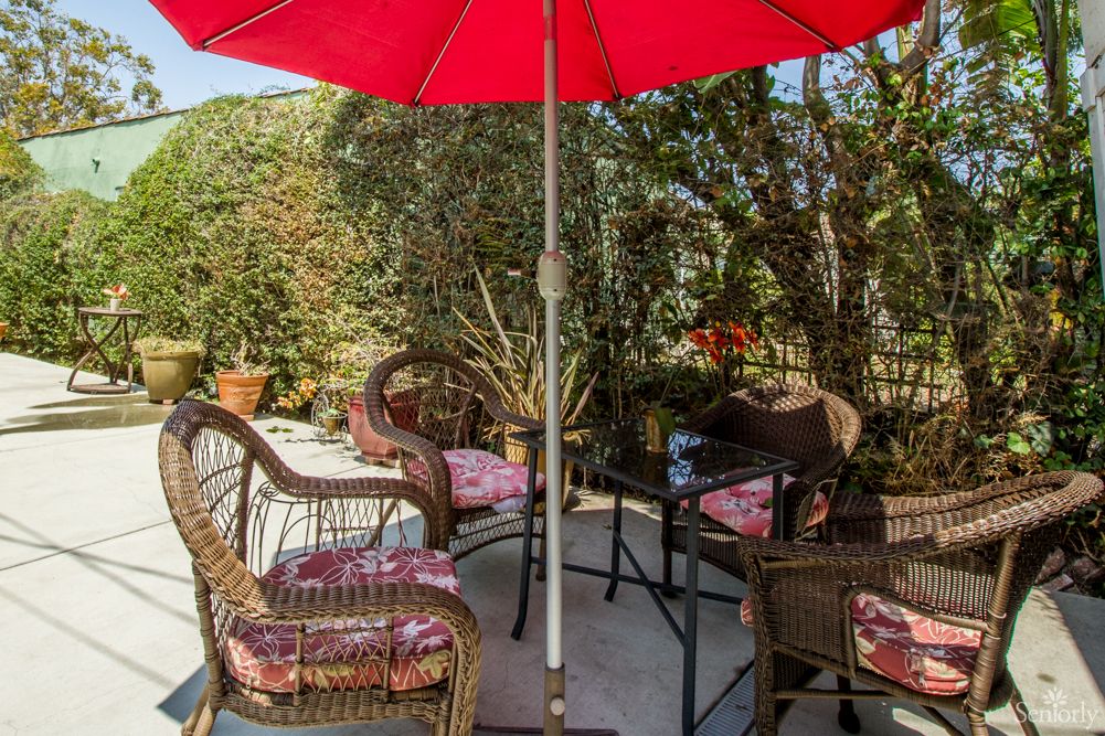 A cozy outdoor patio setup with wicker chairs, a table, and a red umbrella surrounded by lush greenery.