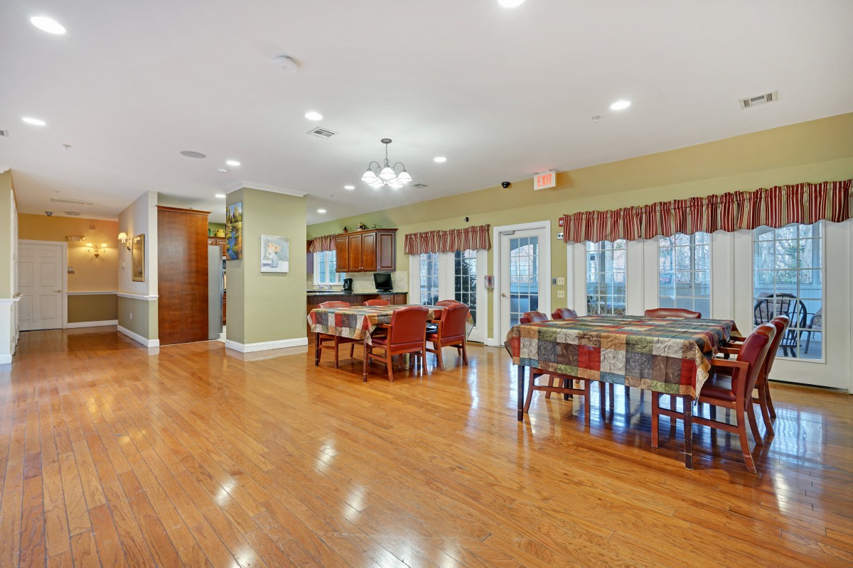 A spacious dining area with hardwood floors, featuring several tables and chairs, adjacent to a kitchen space.
