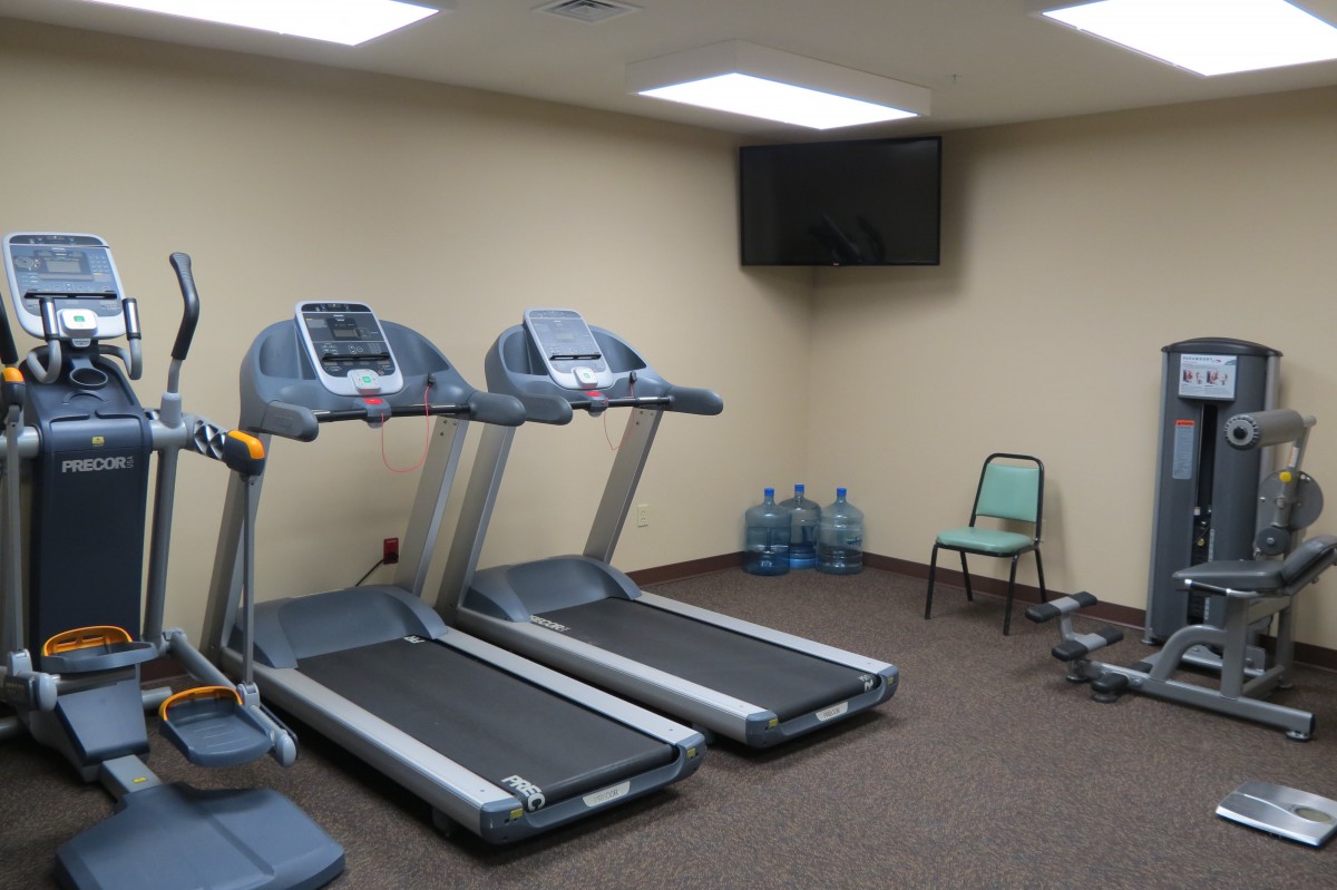 A small gym with two treadmills, a wall-mounted TV, several water bottles, and a chair.