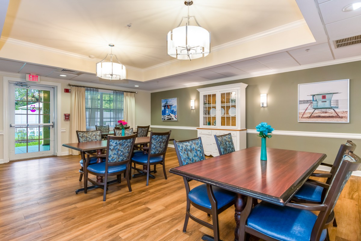 A well-lit dining area with wooden tables and blue chairs, featuring decorative elements such as flowers and artwork on the walls.